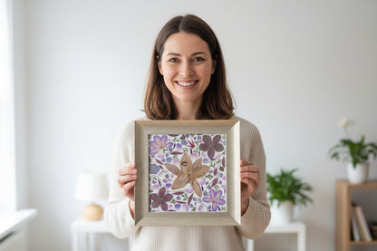 Woman holding a framed floral artwork (contains pressed Amaryllis, purple Cosmos, Ranonkel, leaves) in a room.