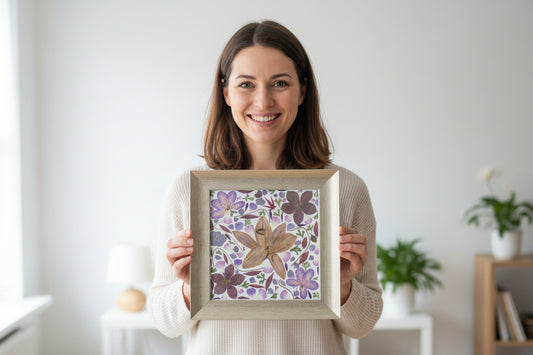 Woman holding a framed floral artwork (contains pressed Amaryllis, purple Cosmos, Ranonkel, leaves) in a room.